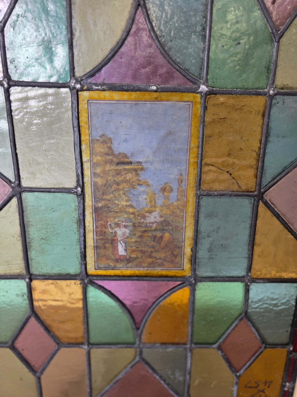 An antique rectangular leaded glass window panel featuring geometric coloured glass panes, bullseye roundels, and a central hand-painted countryside landscape vignette. View III