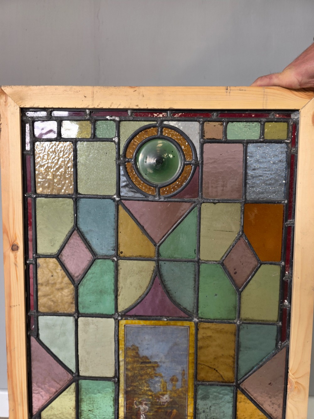 An antique rectangular leaded glass window panel featuring geometric coloured glass panes, bullseye roundels, and a central hand-painted countryside landscape vignette. View IV