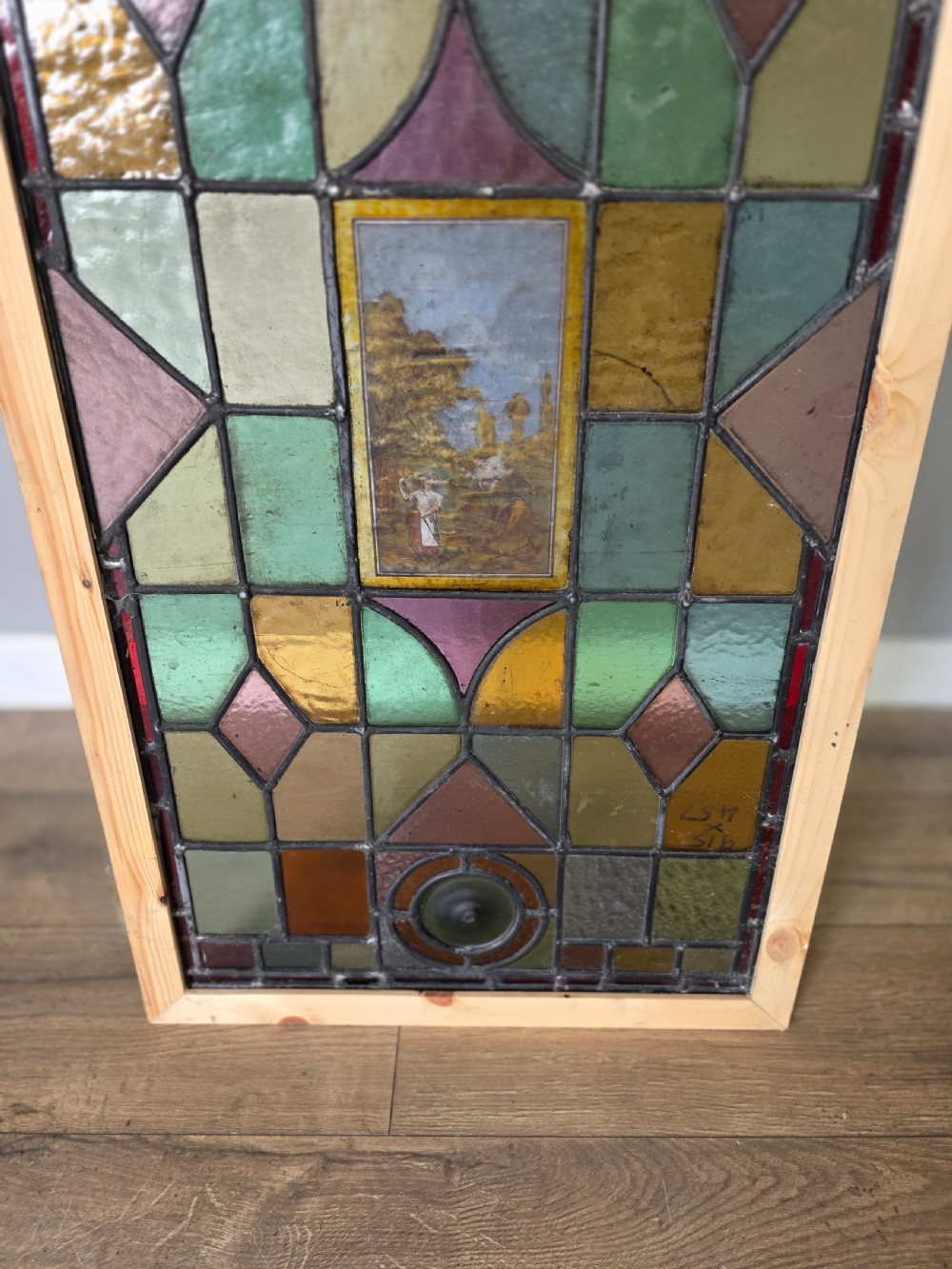 An antique rectangular leaded glass window panel featuring geometric coloured glass panes, bullseye roundels, and a central hand-painted countryside landscape vignette. View V
