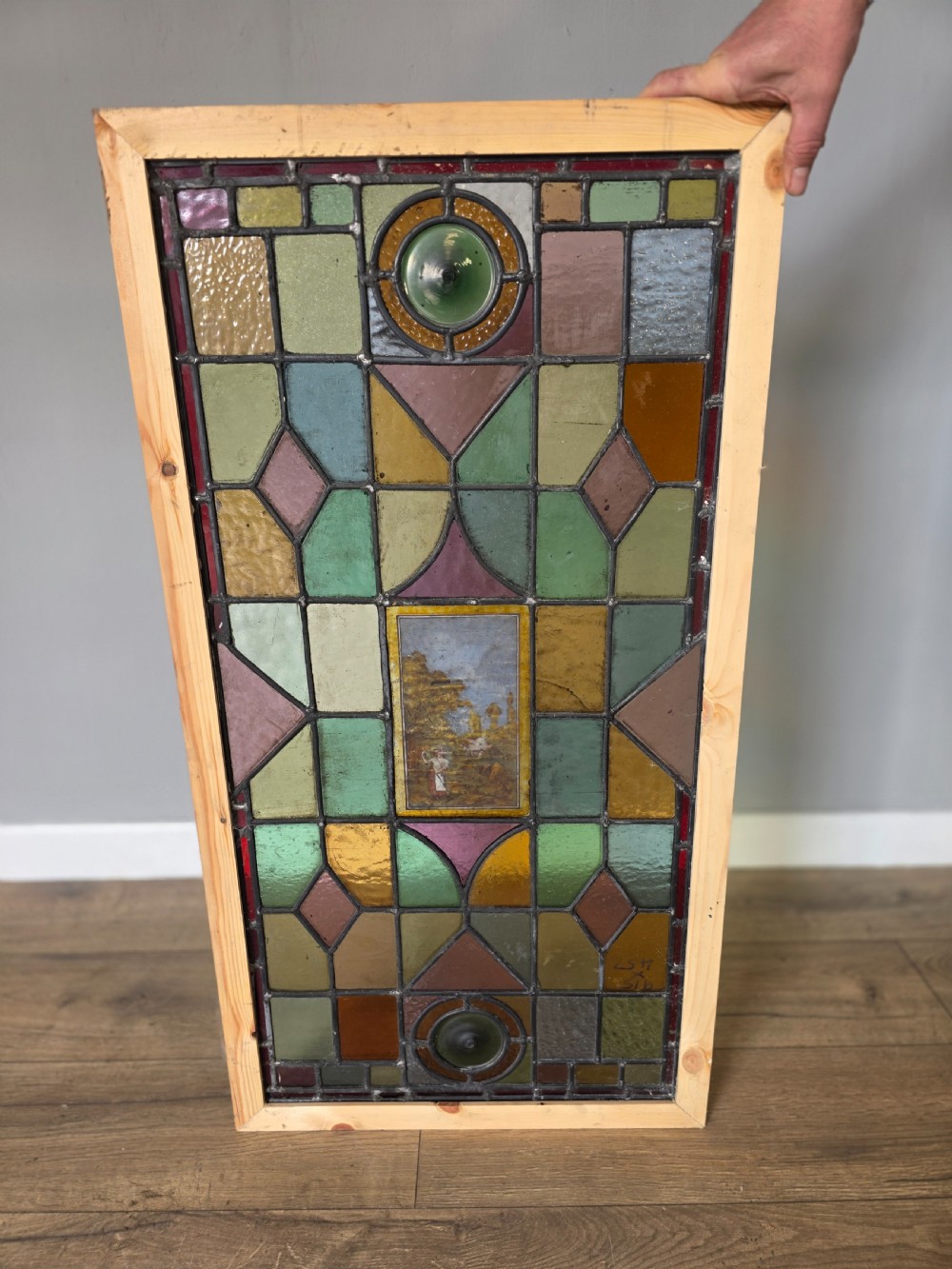 An antique rectangular leaded glass window panel featuring geometric coloured glass panes, bullseye roundels, and a central hand-painted countryside landscape vignette. View VI
