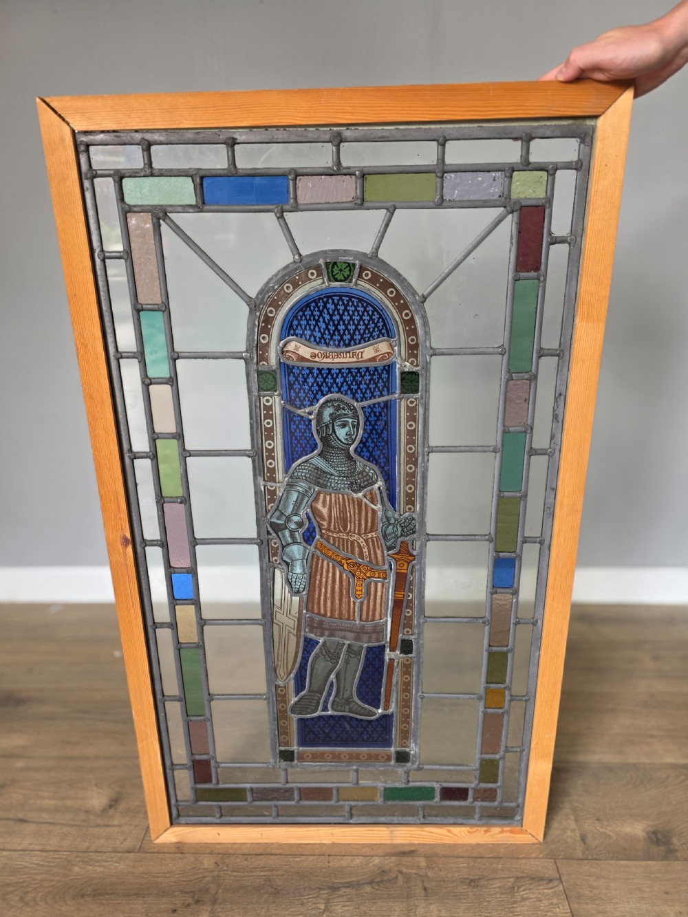 A rectangular leaded glass window panel featuring a central hand-painted figure of a knight in armour against a patterned blue background, encased in a simple pine frame.