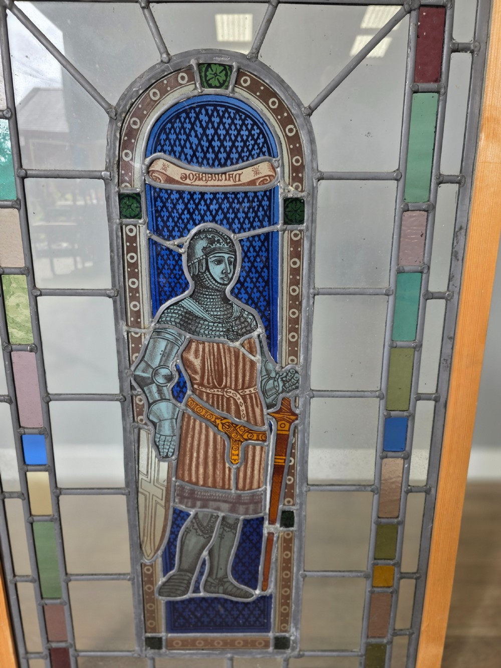 A rectangular leaded glass window panel featuring a central hand-painted figure of a knight in armour against a patterned blue background, encased in a simple pine frame. View II