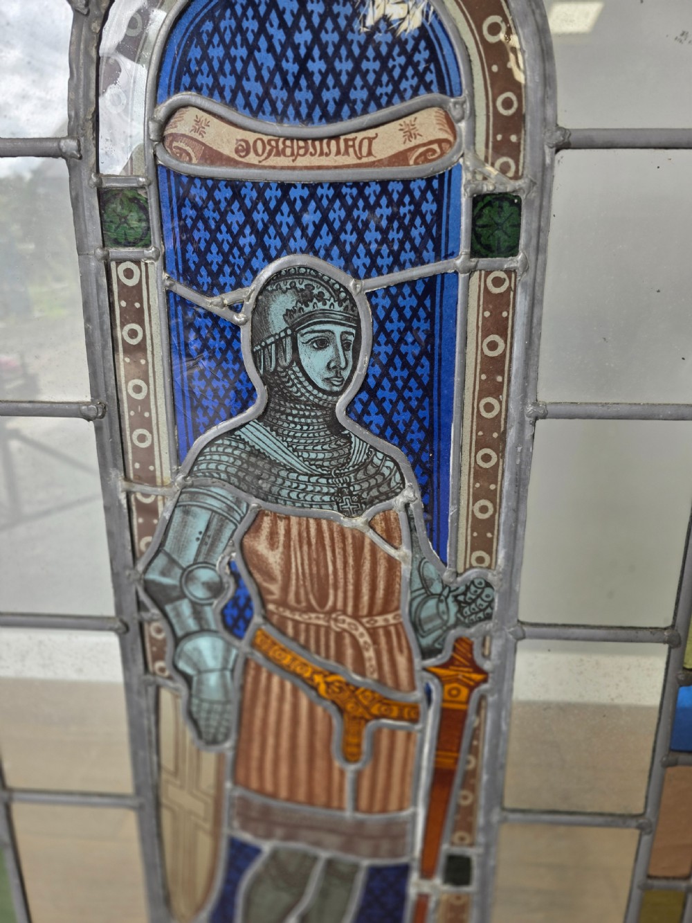 A rectangular leaded glass window panel featuring a central hand-painted figure of a knight in armour against a patterned blue background, encased in a simple pine frame. View III