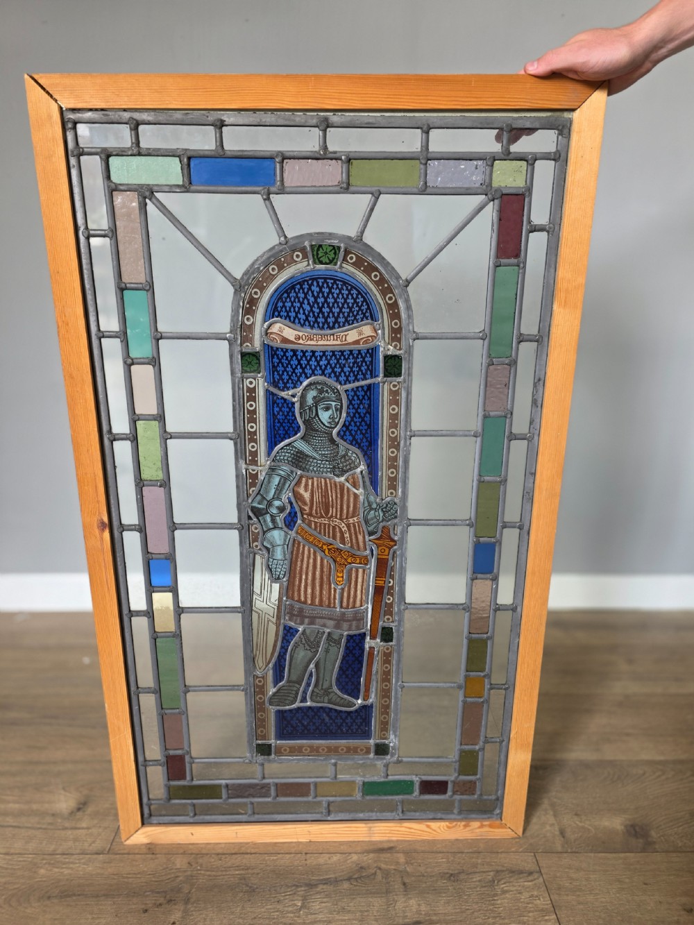 A rectangular leaded glass window panel featuring a central hand-painted figure of a knight in armour against a patterned blue background, encased in a simple pine frame. View IV