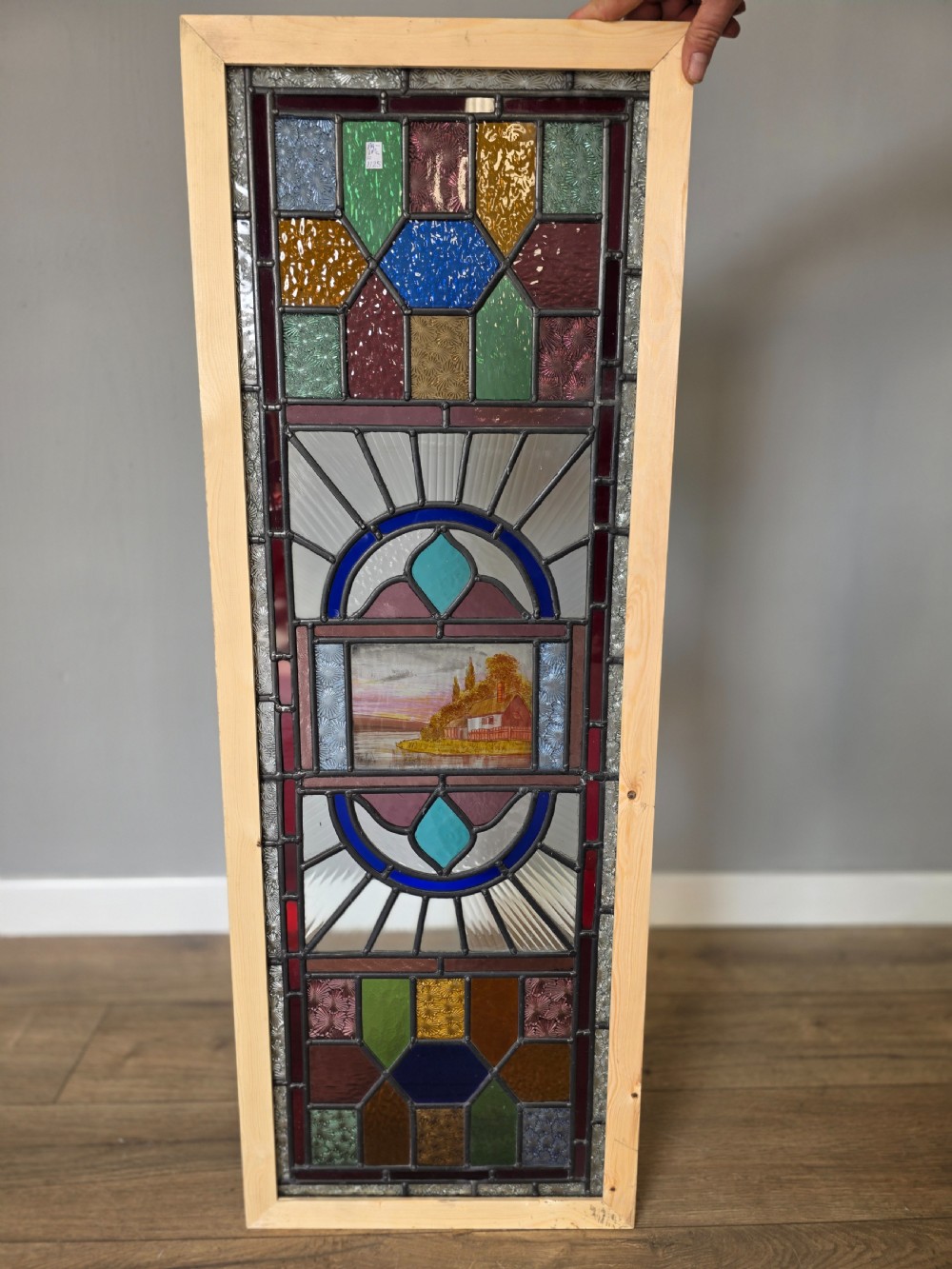 An elongated rectangular leaded glass panel featuring a central hand-painted landscape scene surrounded by geometric coloured glass borders.