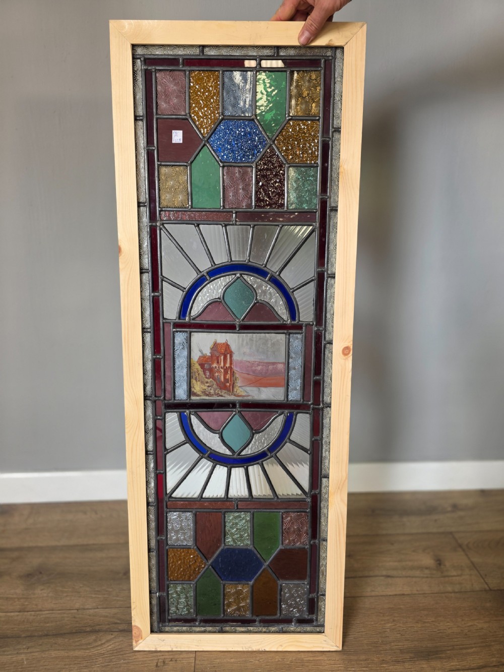 A vertical rectangular leaded glass panel featuring colourful geometric borders, textured glass panes, and a central hand-painted landscape scene.