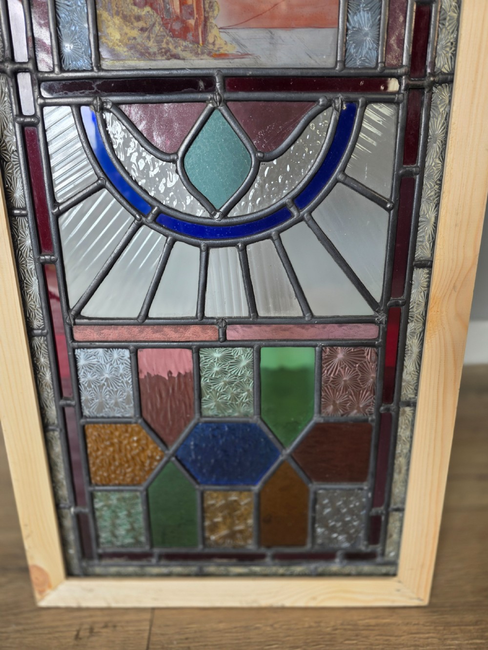A vertical rectangular leaded glass panel featuring colourful geometric borders, textured glass panes, and a central hand-painted landscape scene. View V