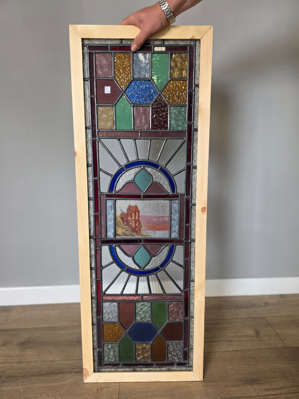 A vertical rectangular leaded glass panel featuring colourful geometric borders, textured glass panes, and a central hand-painted landscape scene. View VI