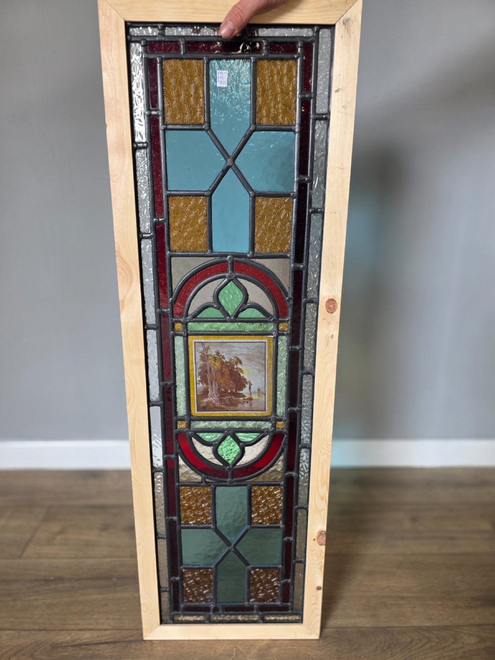 A tall rectangular leaded glass panel set in a light pine wood frame featuring blue, amber, and red geometric glass surrounding a central painted tile depicting a lake and tree landscape.