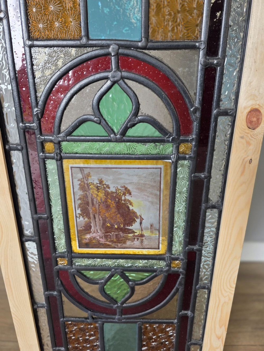 A tall rectangular leaded glass panel set in a light pine wood frame featuring blue, amber, and red geometric glass surrounding a central painted tile depicting a lake and tree landscape. View II