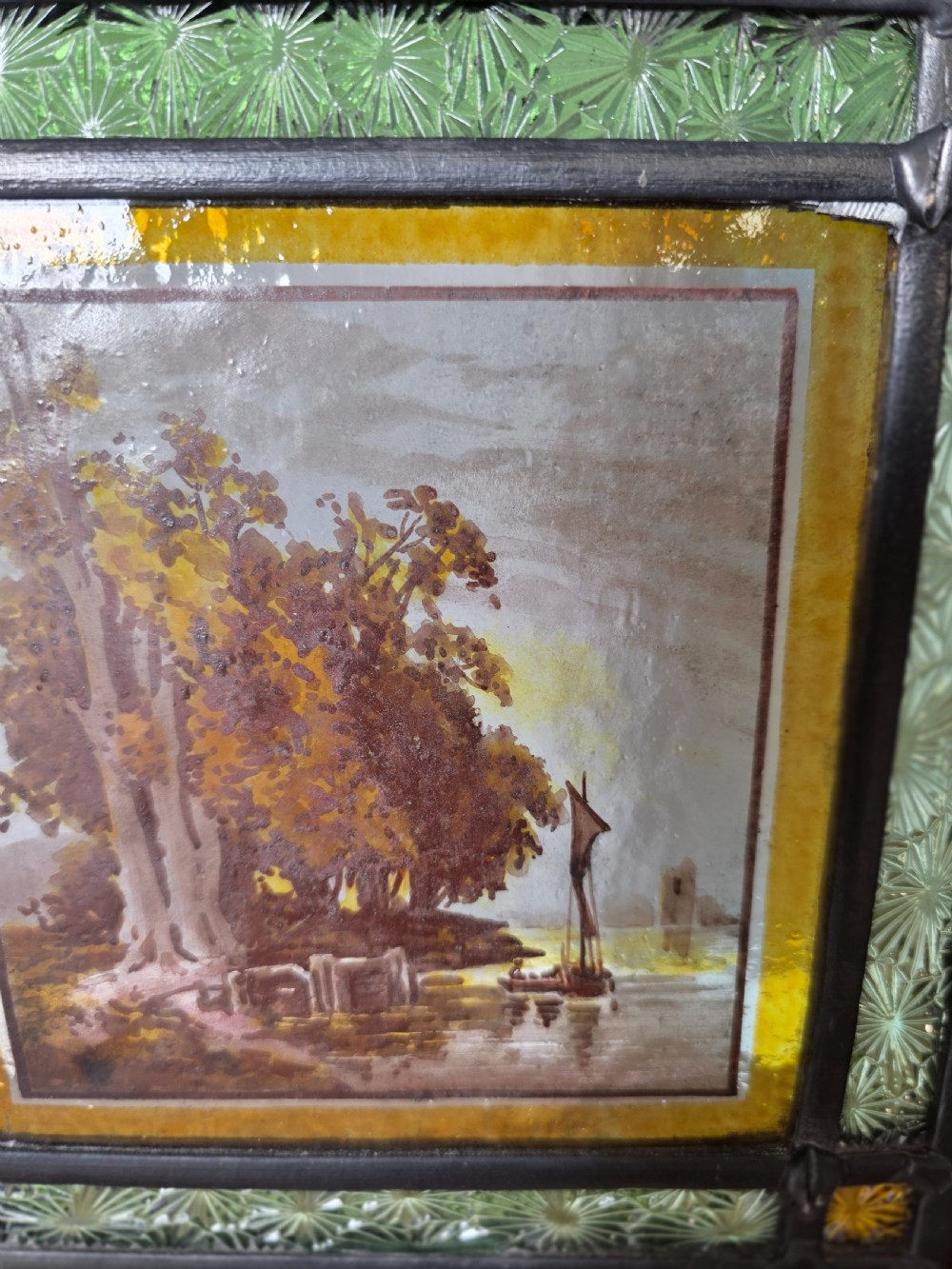A tall rectangular leaded glass panel set in a light pine wood frame featuring blue, amber, and red geometric glass surrounding a central painted tile depicting a lake and tree landscape. View III