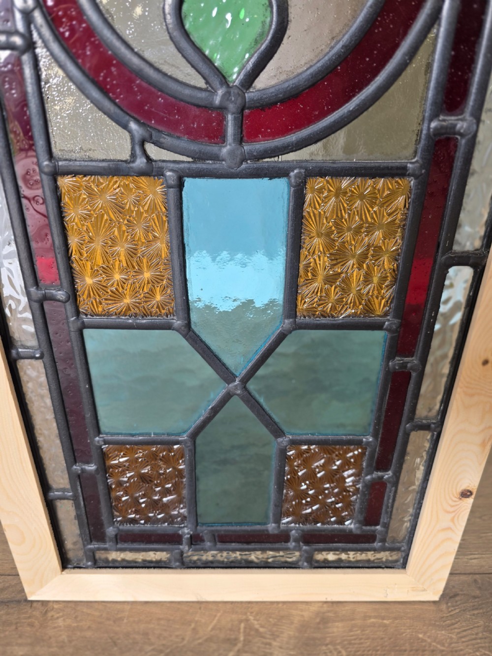 A tall rectangular leaded glass panel set in a light pine wood frame featuring blue, amber, and red geometric glass surrounding a central painted tile depicting a lake and tree landscape. View IV