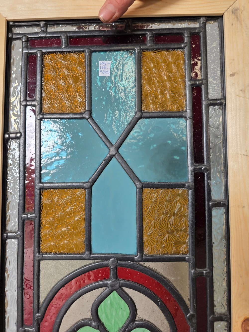 A tall rectangular leaded glass panel set in a light pine wood frame featuring blue, amber, and red geometric glass surrounding a central painted tile depicting a lake and tree landscape. View V