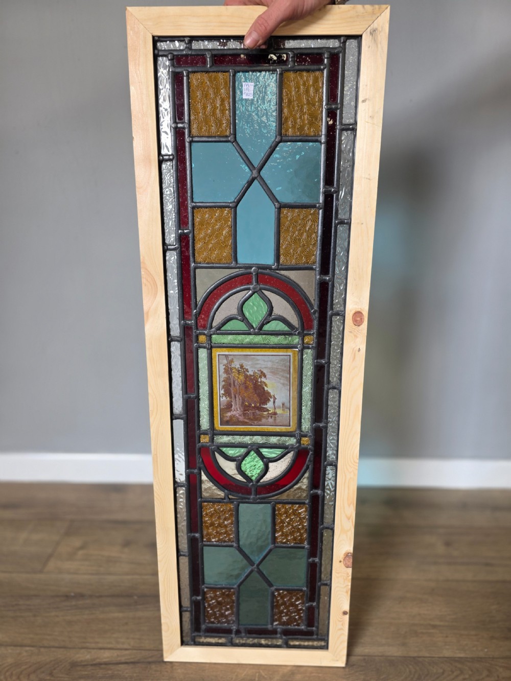 A tall rectangular leaded glass panel set in a light pine wood frame featuring blue, amber, and red geometric glass surrounding a central painted tile depicting a lake and tree landscape. View VI