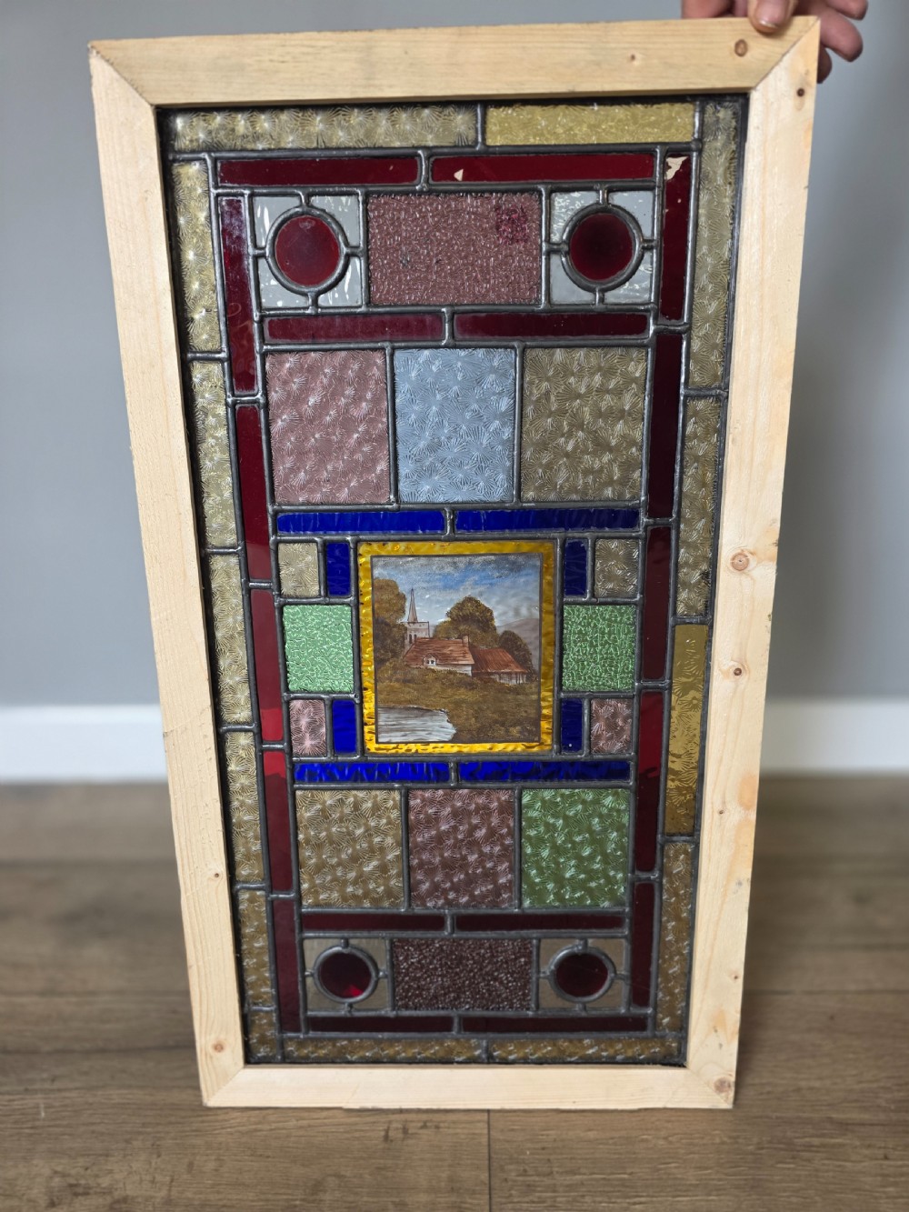 An antique rectangular leaded glass panel featuring colourful geometric textured borders around a central hand-painted countryside landscape scene depicting a church.