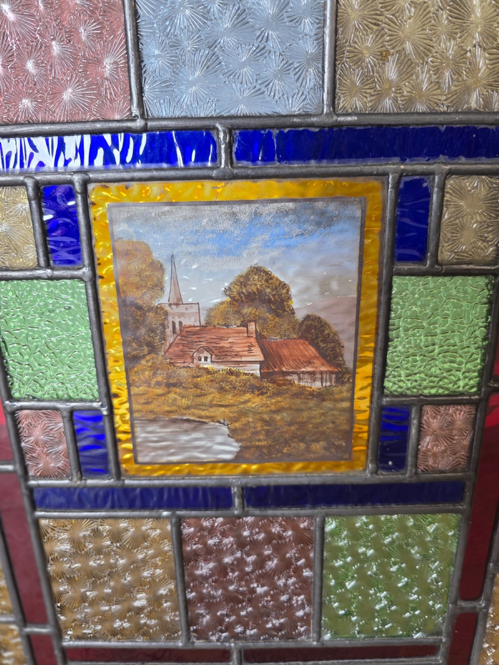 An antique rectangular leaded glass panel featuring colourful geometric textured borders around a central hand-painted countryside landscape scene depicting a church. View II
