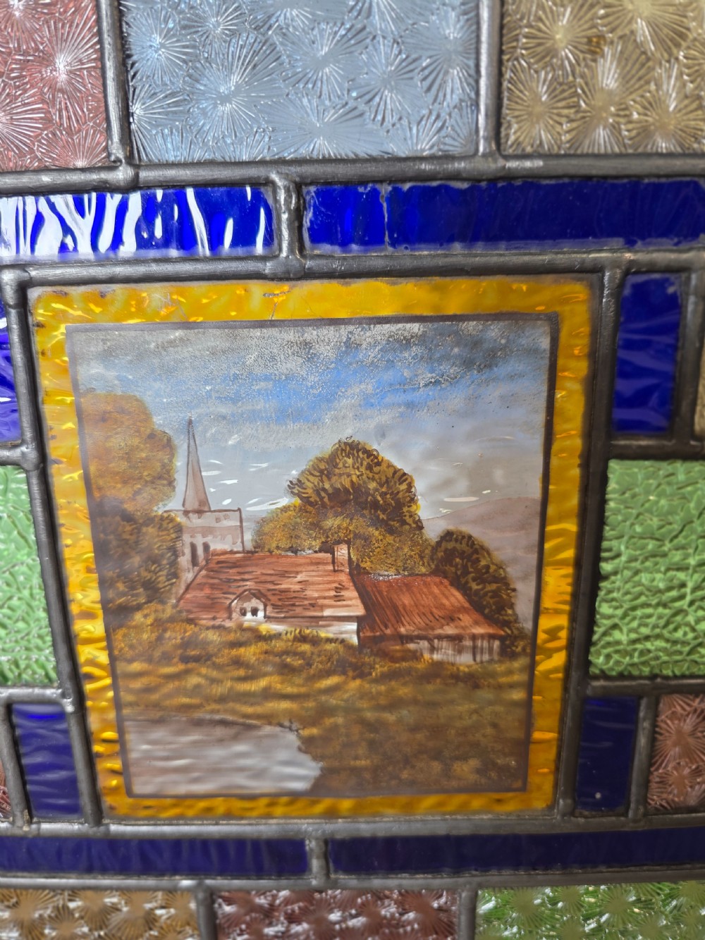 An antique rectangular leaded glass panel featuring colourful geometric textured borders around a central hand-painted countryside landscape scene depicting a church. View III