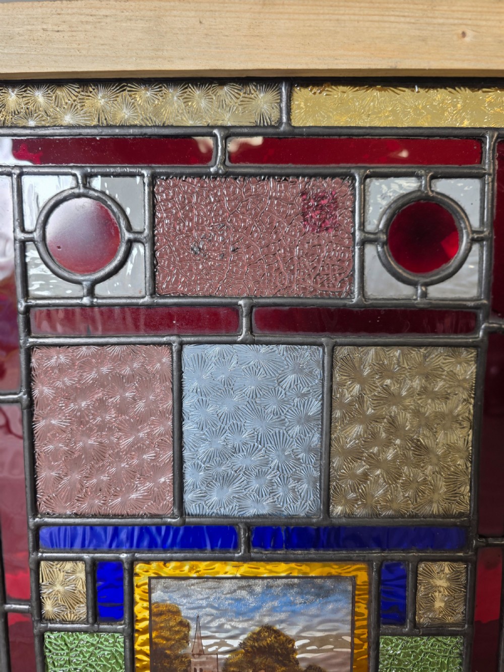 An antique rectangular leaded glass panel featuring colourful geometric textured borders around a central hand-painted countryside landscape scene depicting a church. View IV