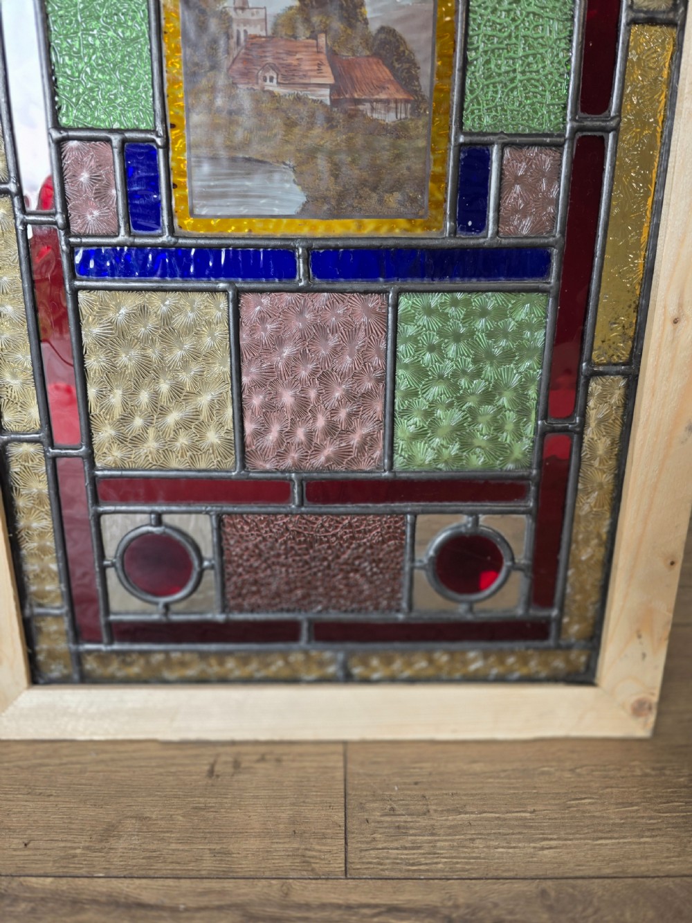 An antique rectangular leaded glass panel featuring colourful geometric textured borders around a central hand-painted countryside landscape scene depicting a church. View V