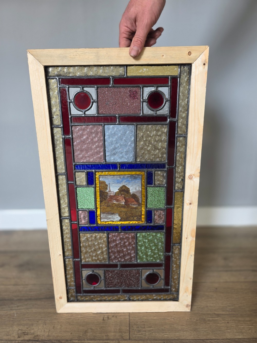 An antique rectangular leaded glass panel featuring colourful geometric textured borders around a central hand-painted countryside landscape scene depicting a church. View VI