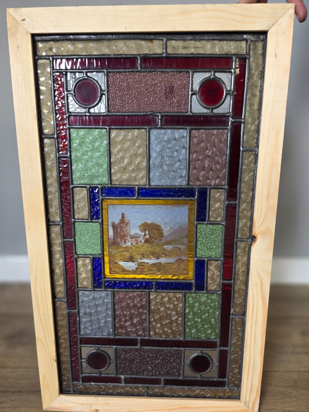 A rectangular Edwardian leaded glass window panel set in a light pine frame, featuring a central hand-painted landscape with a ruined castle surrounded by geometric textured coloured glass.