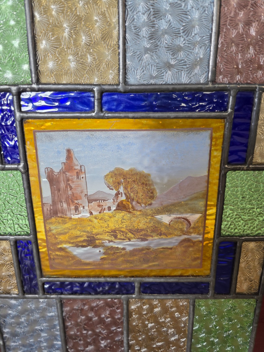 A rectangular Edwardian leaded glass window panel set in a light pine frame, featuring a central hand-painted landscape with a ruined castle surrounded by geometric textured coloured glass. View II