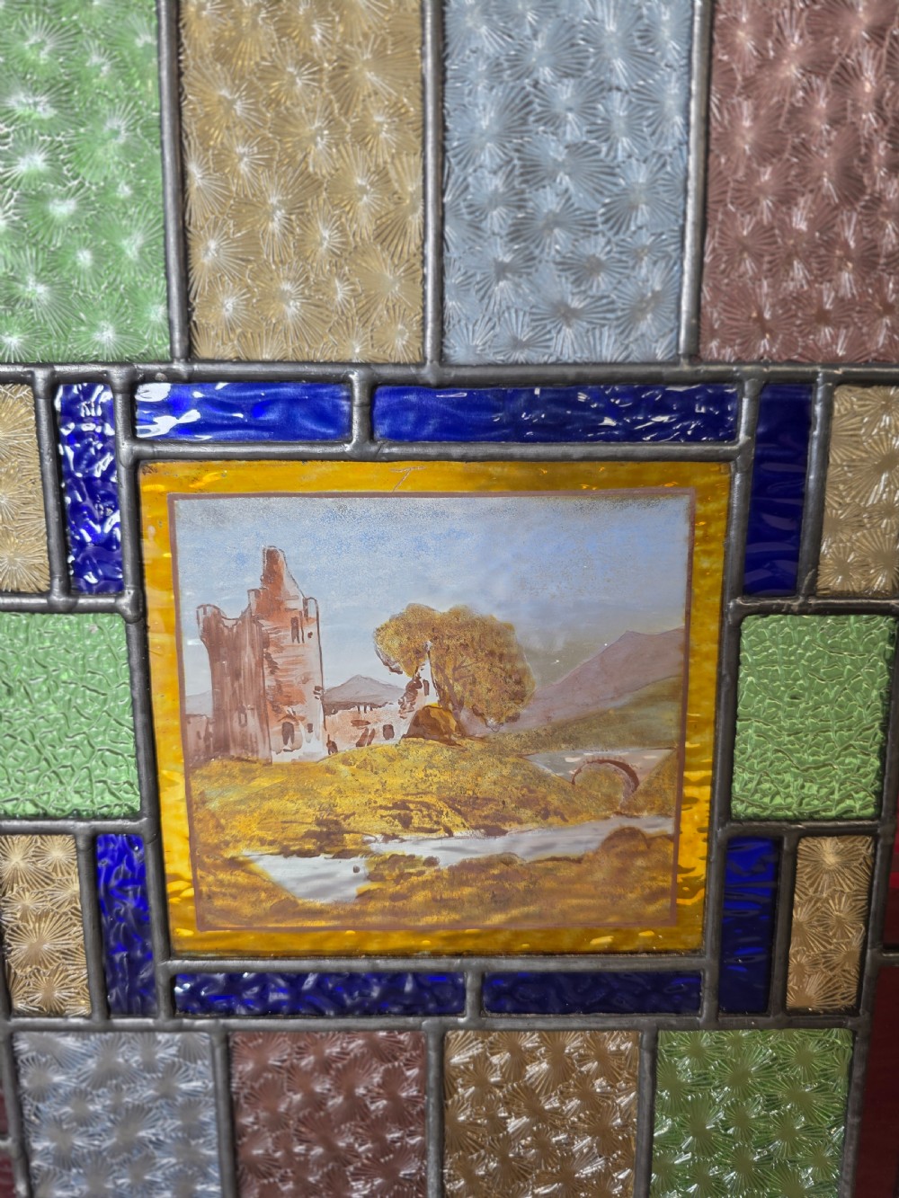 A rectangular Edwardian leaded glass window panel set in a light pine frame, featuring a central hand-painted landscape with a ruined castle surrounded by geometric textured coloured glass. View III