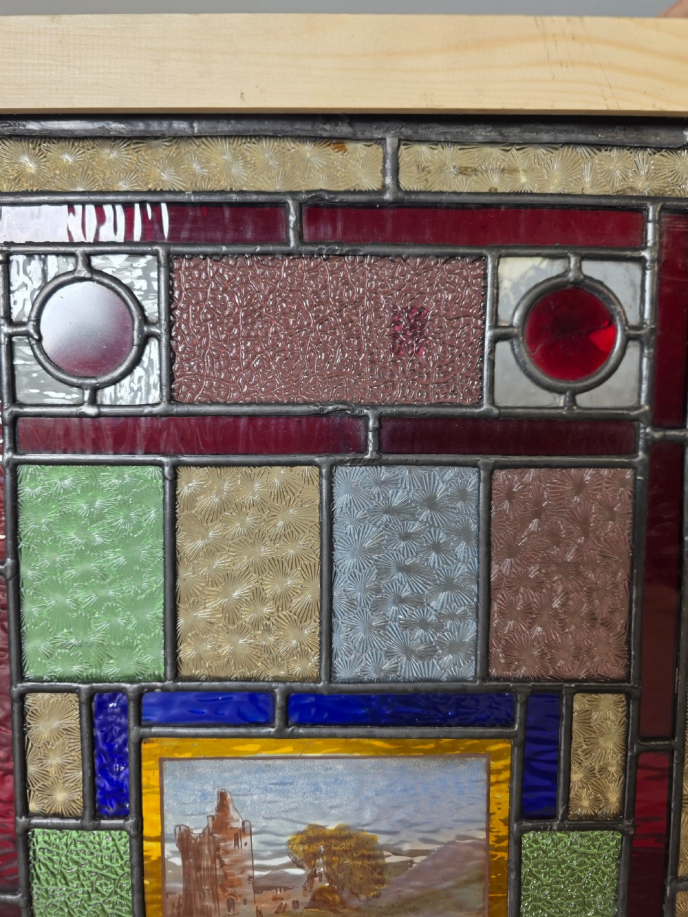 A rectangular Edwardian leaded glass window panel set in a light pine frame, featuring a central hand-painted landscape with a ruined castle surrounded by geometric textured coloured glass. View IV