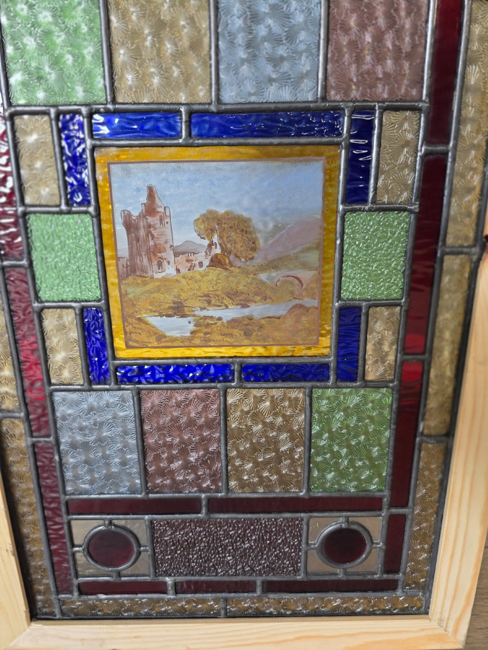 A rectangular Edwardian leaded glass window panel set in a light pine frame, featuring a central hand-painted landscape with a ruined castle surrounded by geometric textured coloured glass. View V