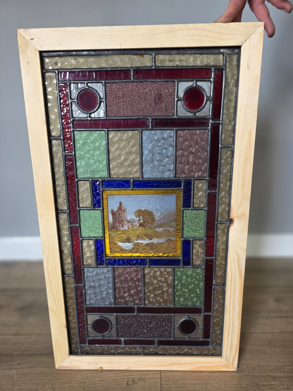 A rectangular Edwardian leaded glass window panel set in a light pine frame, featuring a central hand-painted landscape with a ruined castle surrounded by geometric textured coloured glass. View VI