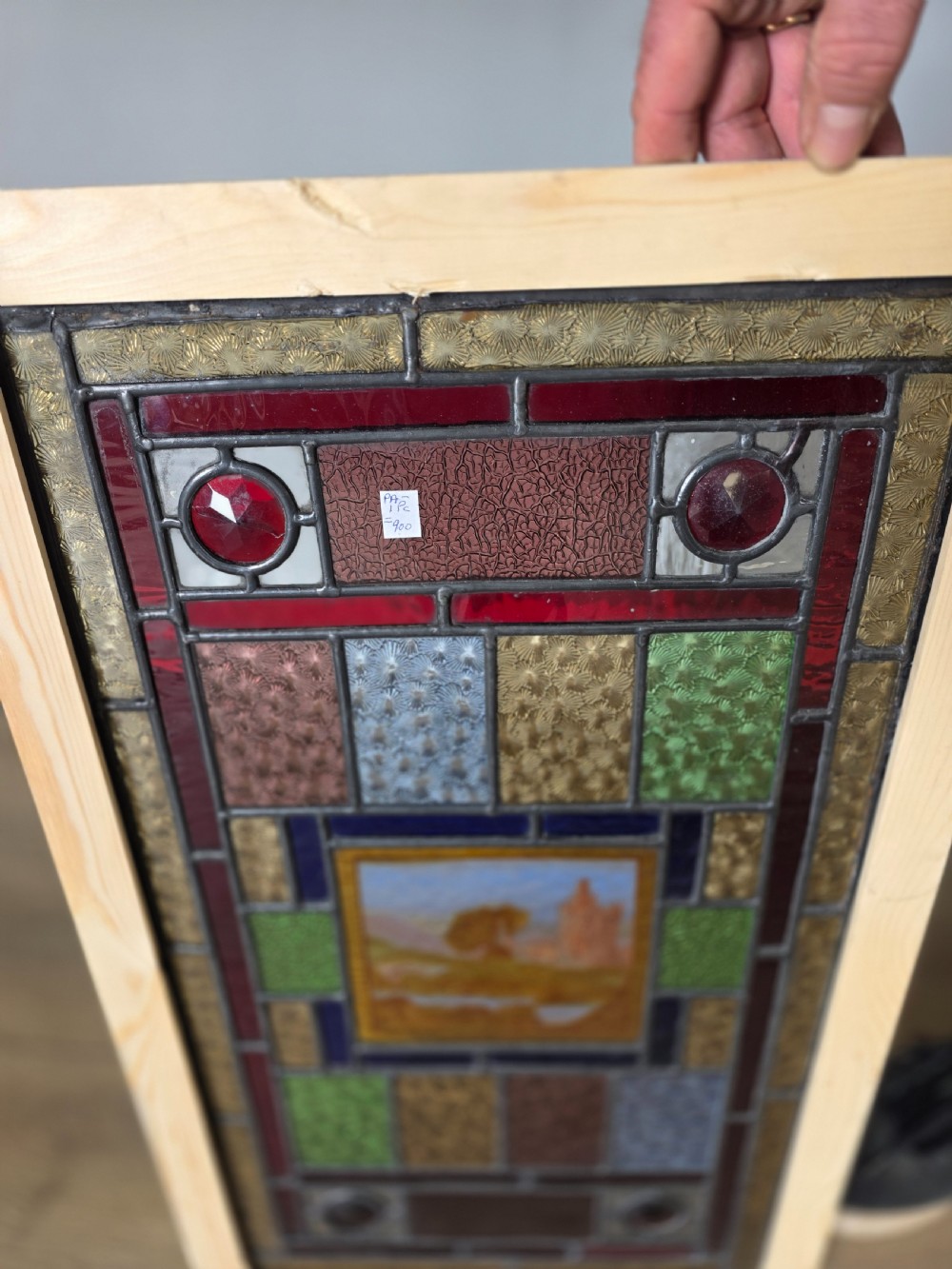 A rectangular Edwardian leaded glass window panel set in a light pine frame, featuring a central hand-painted landscape with a ruined castle surrounded by geometric textured coloured glass. View VII