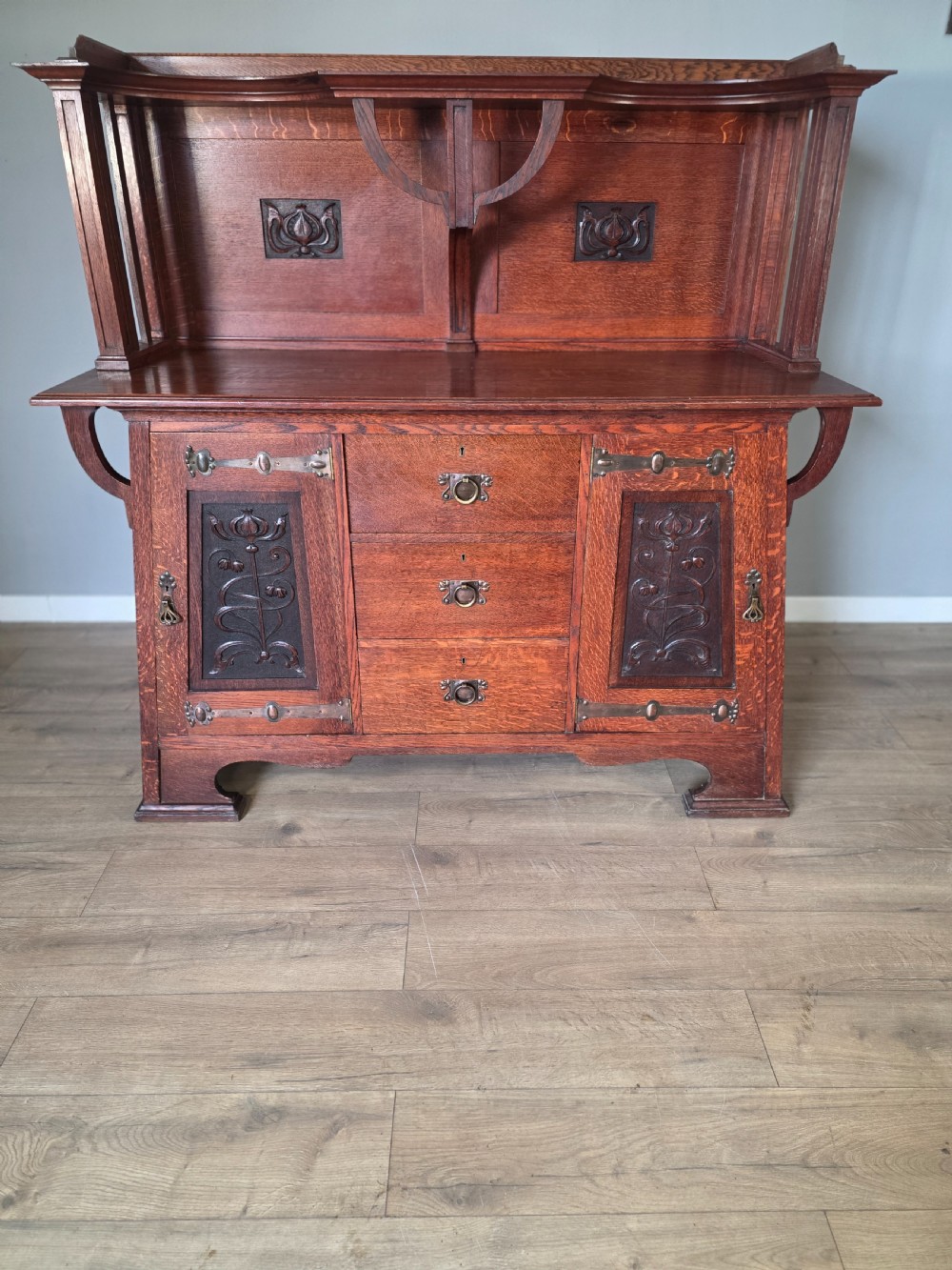 An Arts and Crafts quarter-sawn oak sideboard featuring hand-carved floral panels, copper strap hinges, central drawers, and a raised open shelf superstructure.