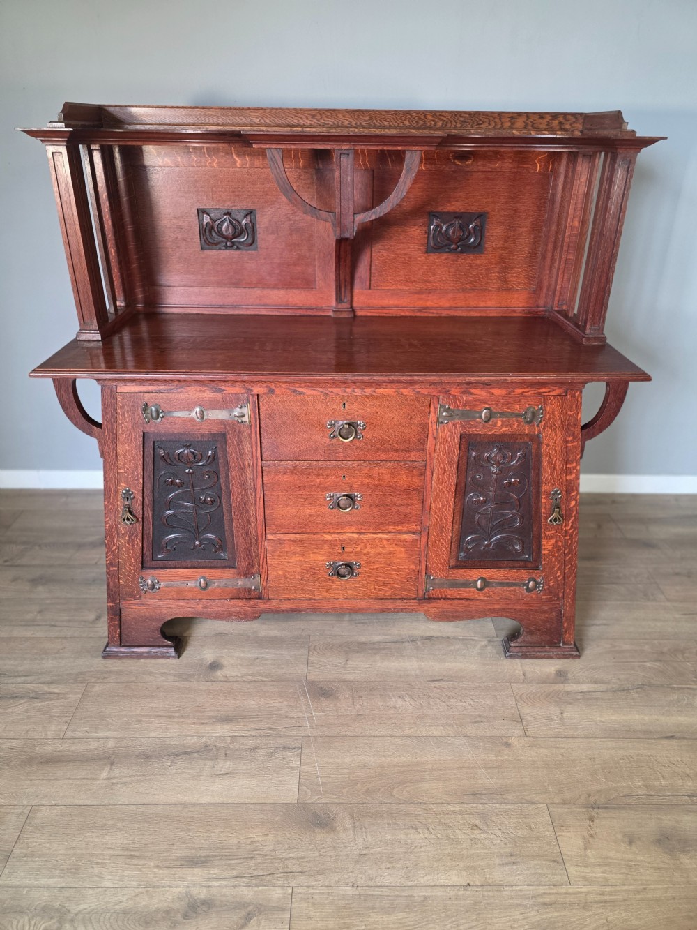 An Arts and Crafts quarter-sawn oak sideboard featuring hand-carved floral panels, copper strap hinges, central drawers, and a raised open shelf superstructure. View II