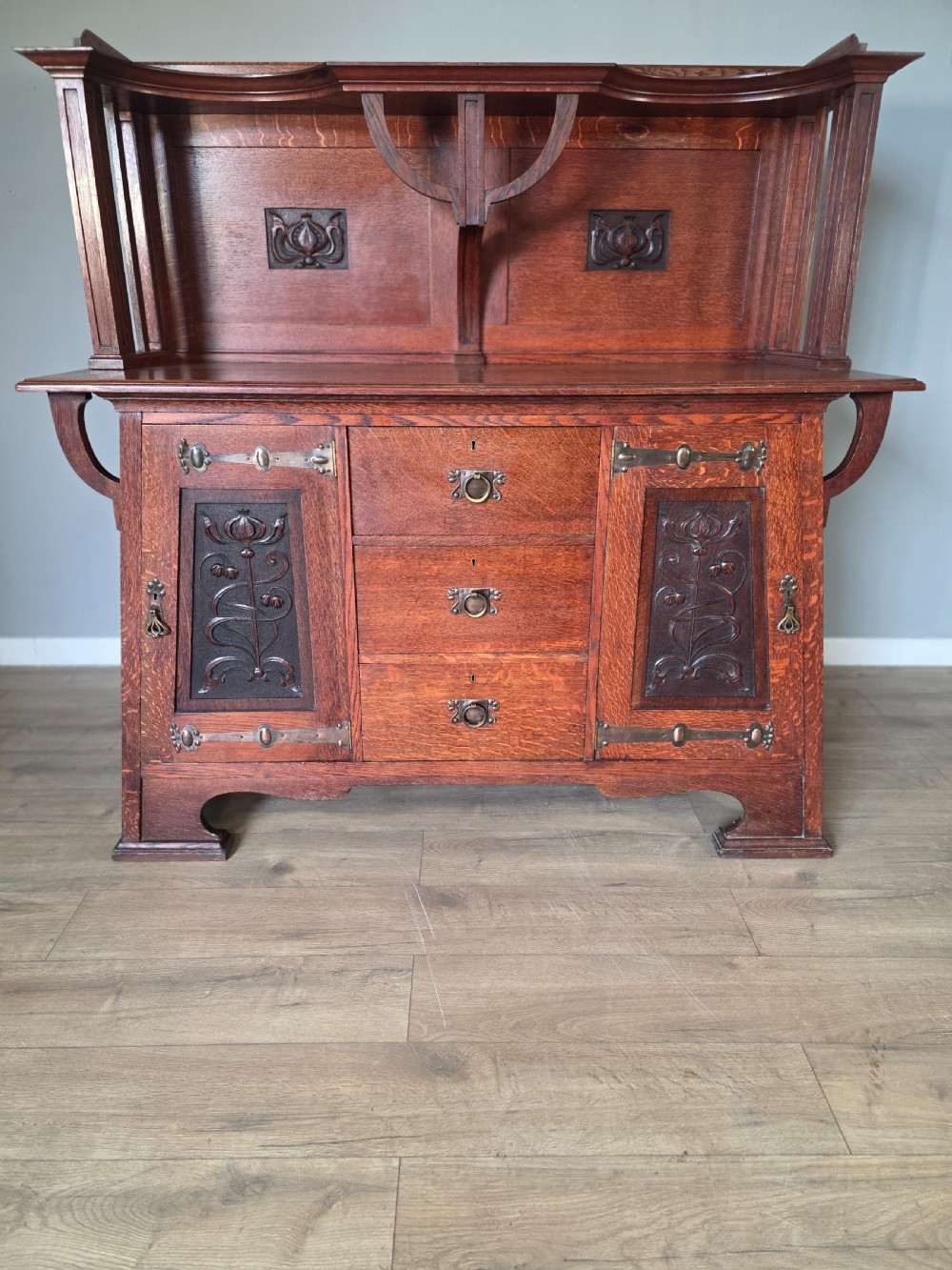 An Arts and Crafts quarter-sawn oak sideboard featuring hand-carved floral panels, copper strap hinges, central drawers, and a raised open shelf superstructure. View III