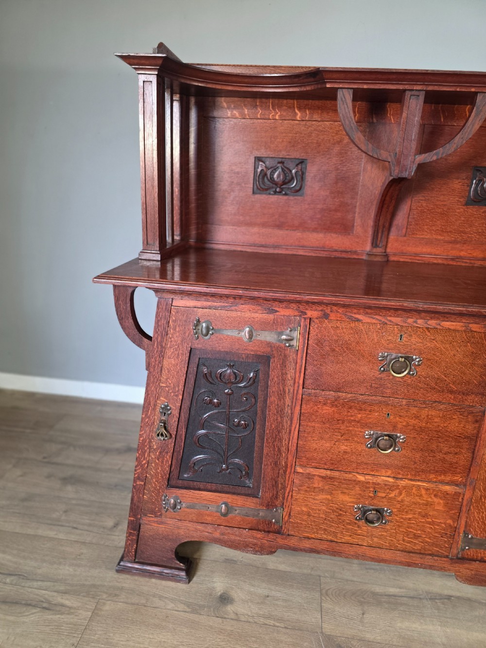 An Arts and Crafts quarter-sawn oak sideboard featuring hand-carved floral panels, copper strap hinges, central drawers, and a raised open shelf superstructure. View IV