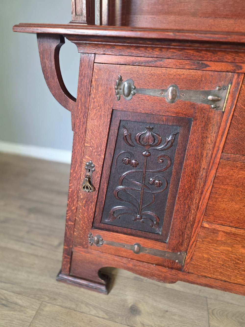 An Arts and Crafts quarter-sawn oak sideboard featuring hand-carved floral panels, copper strap hinges, central drawers, and a raised open shelf superstructure. View V