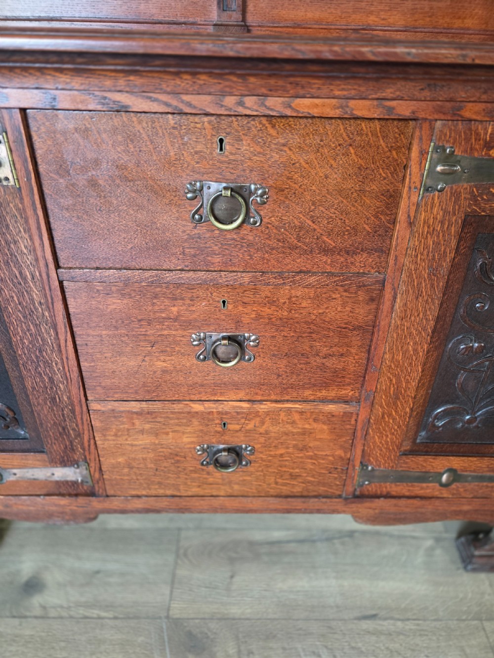 An Arts and Crafts quarter-sawn oak sideboard featuring hand-carved floral panels, copper strap hinges, central drawers, and a raised open shelf superstructure. View VI