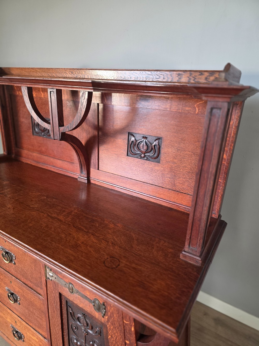 An Arts and Crafts quarter-sawn oak sideboard featuring hand-carved floral panels, copper strap hinges, central drawers, and a raised open shelf superstructure. View VIII