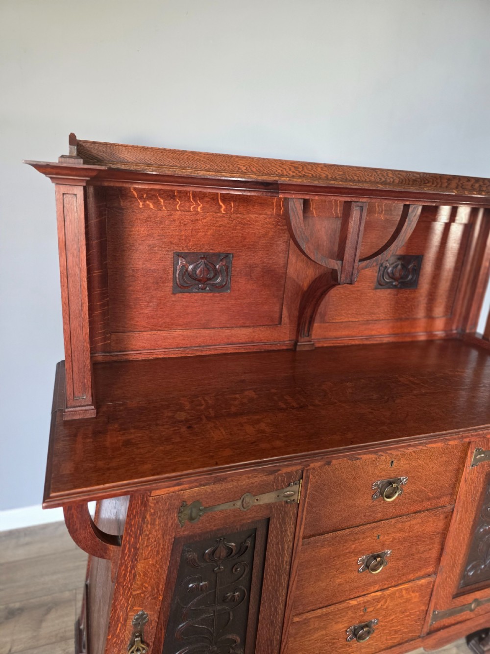 An Arts and Crafts quarter-sawn oak sideboard featuring hand-carved floral panels, copper strap hinges, central drawers, and a raised open shelf superstructure. View IX