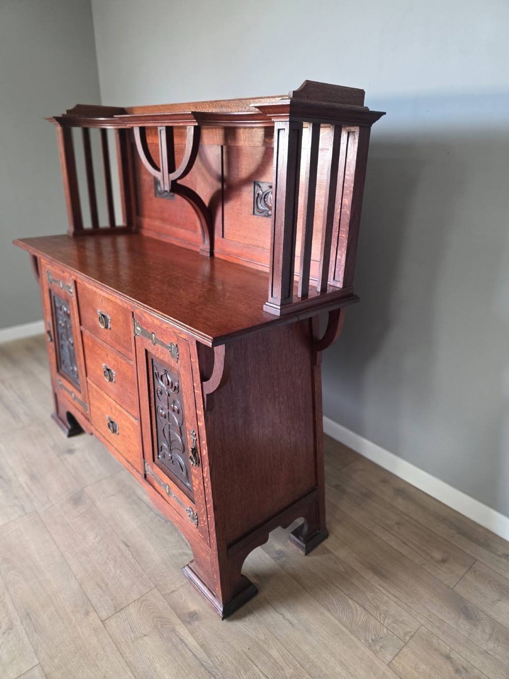 An Arts and Crafts quarter-sawn oak sideboard featuring hand-carved floral panels, copper strap hinges, central drawers, and a raised open shelf superstructure. View XI