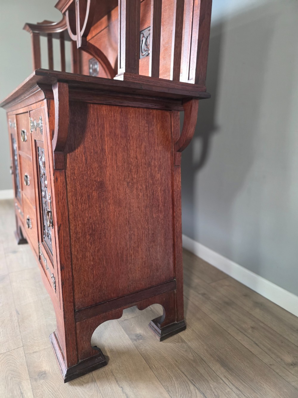 An Arts and Crafts quarter-sawn oak sideboard featuring hand-carved floral panels, copper strap hinges, central drawers, and a raised open shelf superstructure. View XII