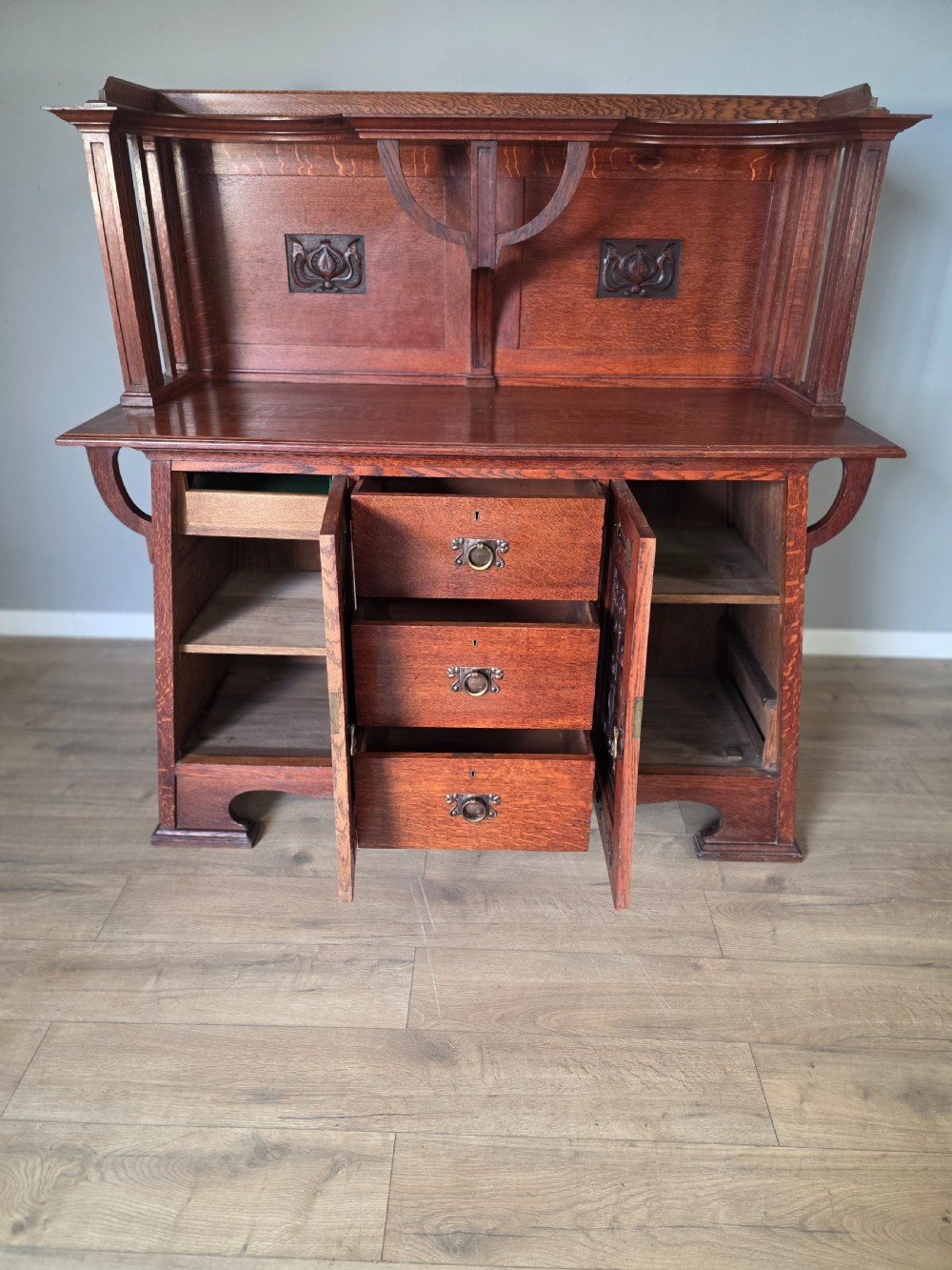 An Arts and Crafts quarter-sawn oak sideboard featuring hand-carved floral panels, copper strap hinges, central drawers, and a raised open shelf superstructure. View XIII