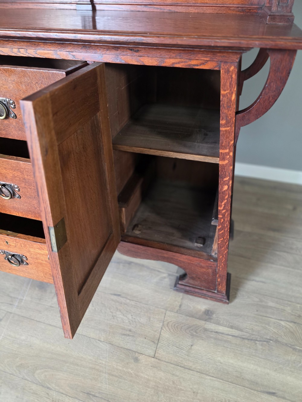 An Arts and Crafts quarter-sawn oak sideboard featuring hand-carved floral panels, copper strap hinges, central drawers, and a raised open shelf superstructure. View XIV