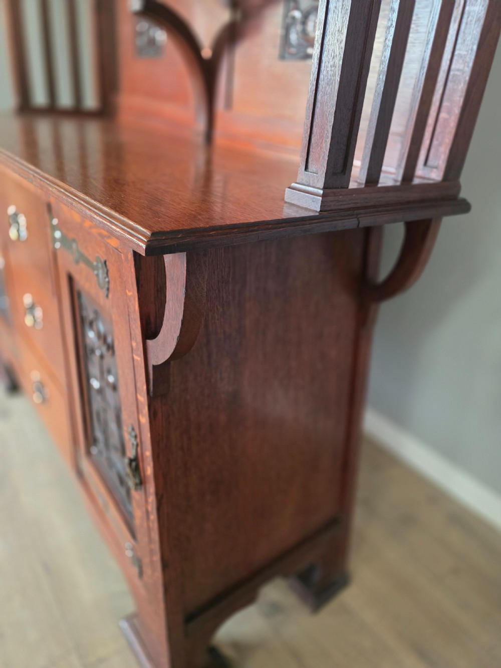An Arts and Crafts quarter-sawn oak sideboard featuring hand-carved floral panels, copper strap hinges, central drawers, and a raised open shelf superstructure. View XVIII