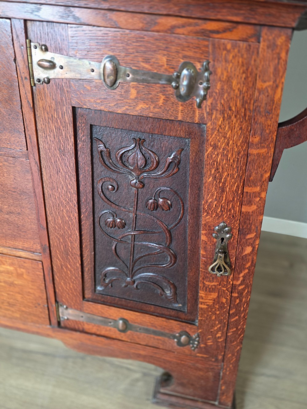 An Arts and Crafts quarter-sawn oak sideboard featuring hand-carved floral panels, copper strap hinges, central drawers, and a raised open shelf superstructure. View XIX