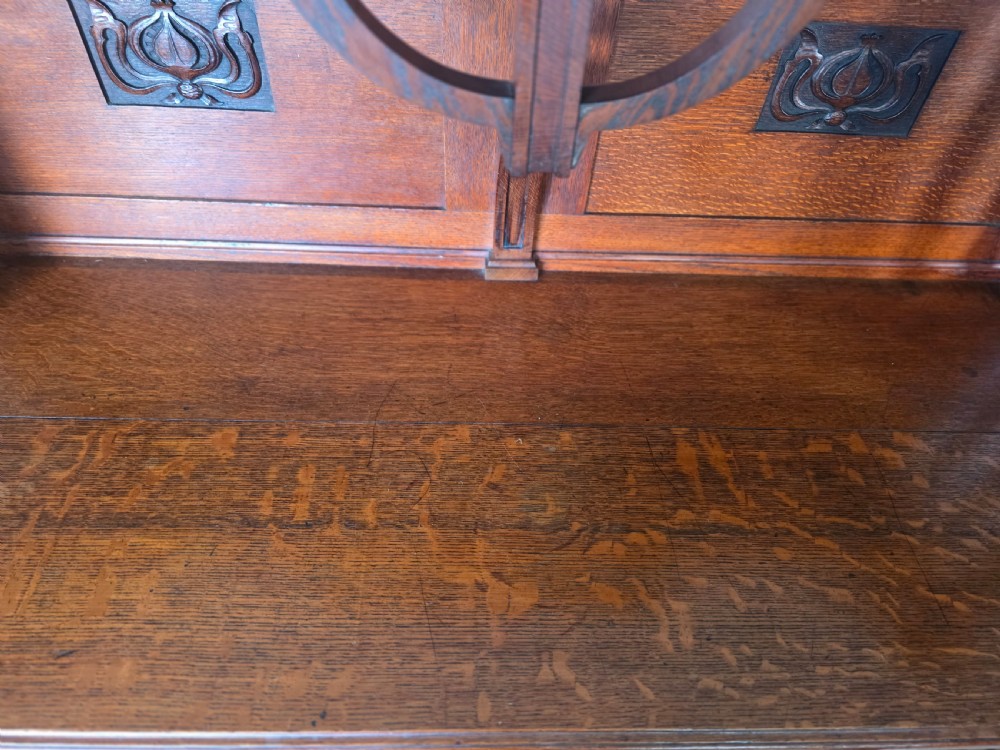 An Arts and Crafts quarter-sawn oak sideboard featuring hand-carved floral panels, copper strap hinges, central drawers, and a raised open shelf superstructure. View XXI