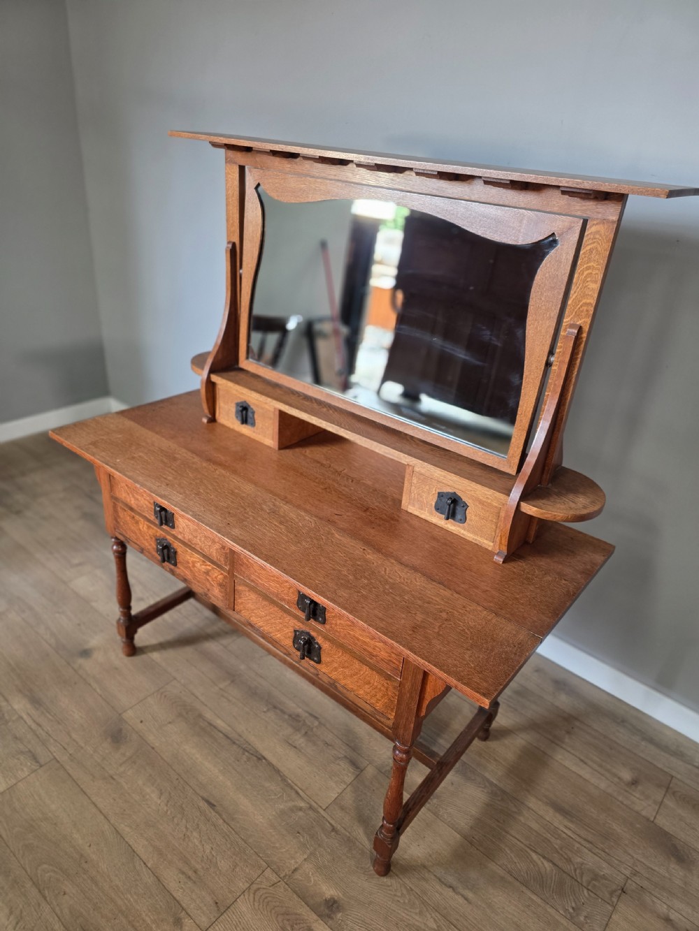 An Arts and Crafts solid oak dressing table featuring an upper mirror superstructure with side trinket drawers above a lower four-drawer chest with dark metal handles. View III