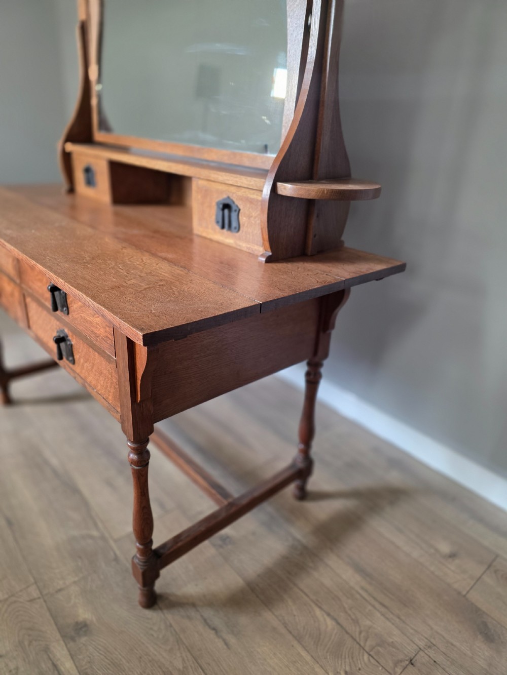 An Arts and Crafts solid oak dressing table featuring an upper mirror superstructure with side trinket drawers above a lower four-drawer chest with dark metal handles. View XIII