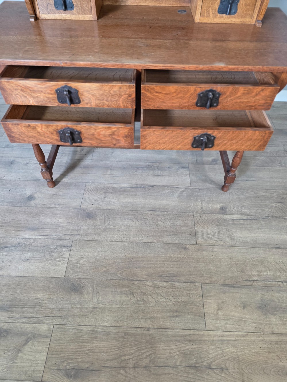 An Arts and Crafts solid oak dressing table featuring an upper mirror superstructure with side trinket drawers above a lower four-drawer chest with dark metal handles. View XV