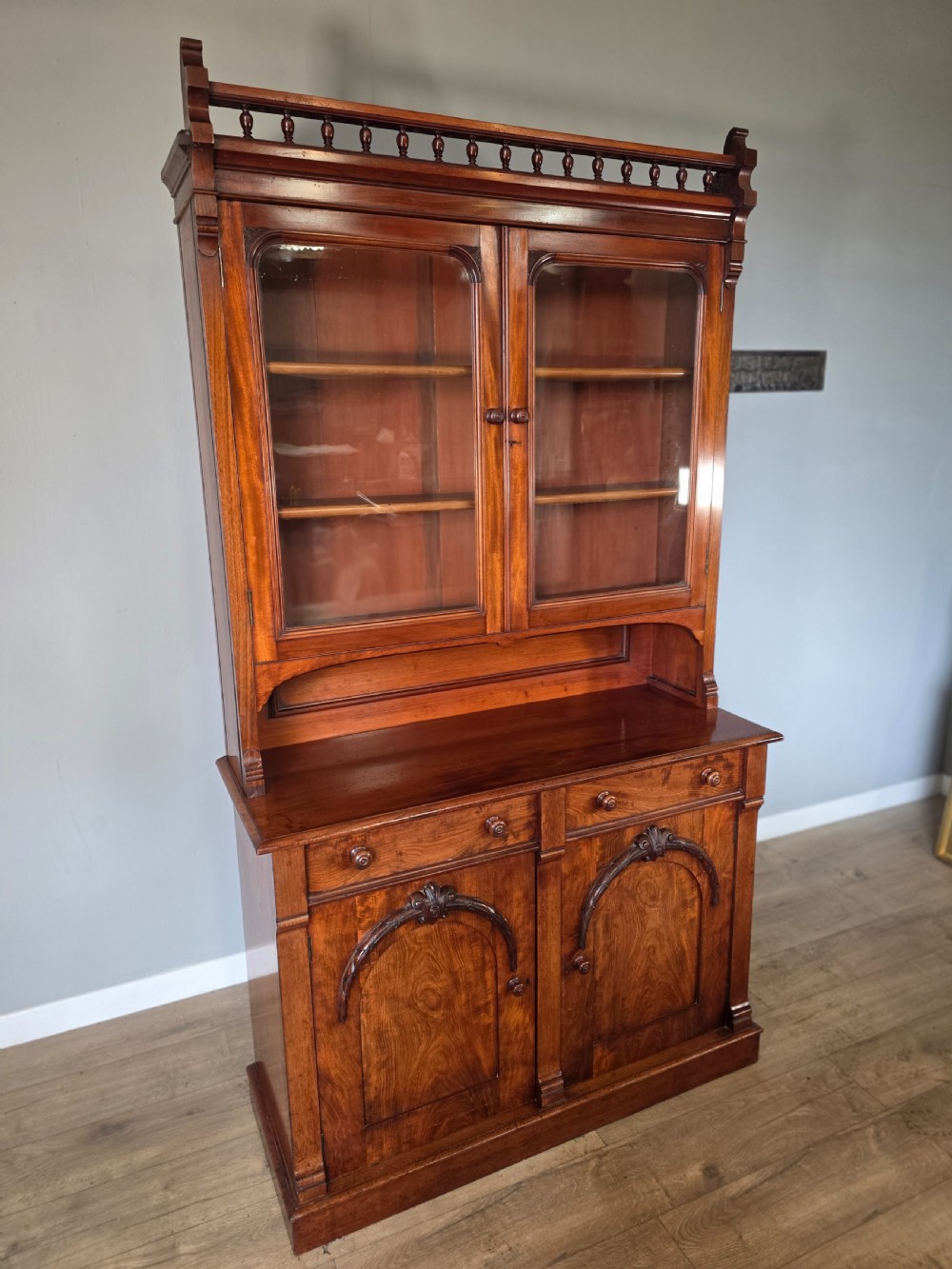 A Victorian mahogany glazed bookcase featuring a top spindle gallery, two glass cabinet doors, two drawers, and a lower double door cupboard on a plinth base. View V